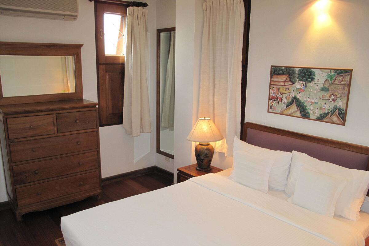 A hotel room with a wooden dresser, a bed with white linens, a nightstand with a lamp, and a wall painting.