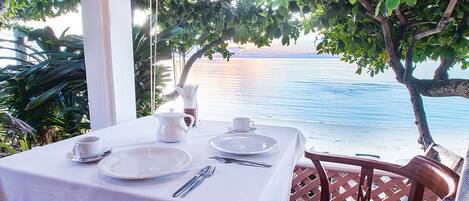 A table set for two with white tablecloth, plates, cutlery, and a white pitcher, overlooking a beach with a clear view of the sea and palm trees.