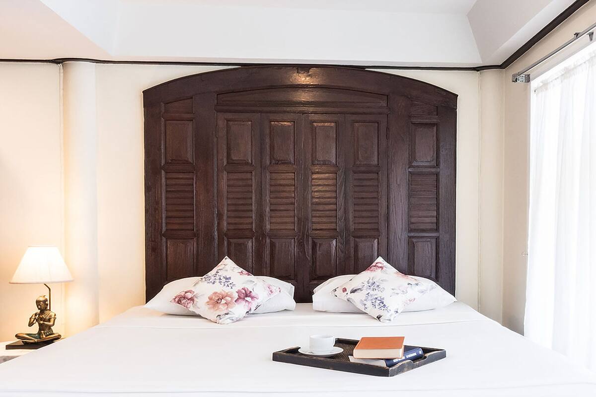 A hotel room with a large dark wooden headboard, a bed with floral pillows, a bedside table with a lamp, and a tray with a book and a cup.