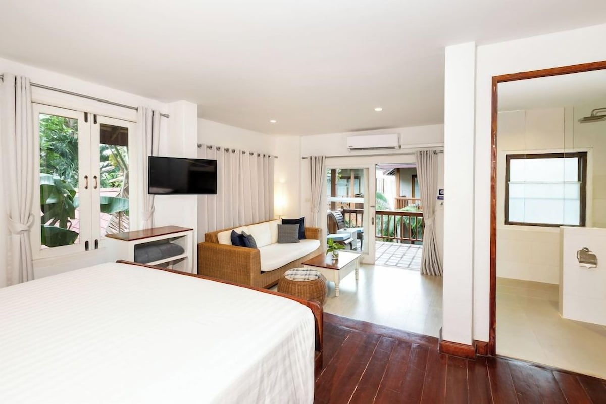 A hotel room with a bed, a sofa, a TV, a balcony with a table and chairs, and an air conditioning unit.