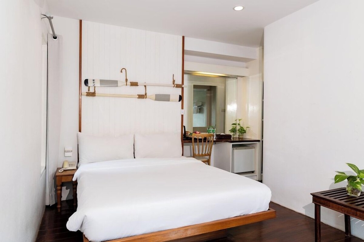 A bedroom with a bed, a desk, a chair, a small table with a plant, and a mirror.