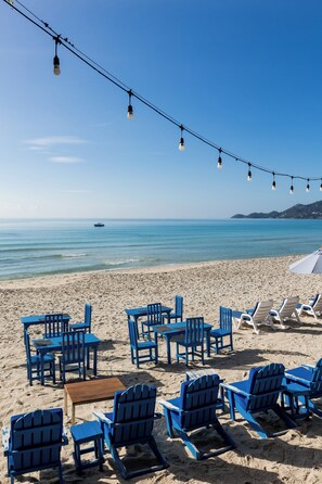 A beachside seating area with tables and chairs, a string of lights, and a view of the sea. - Malibu Beach Resort Samui (Koh Samui)