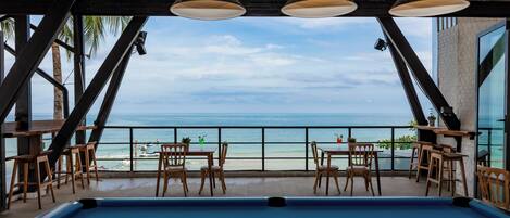 A pool table with balls, wooden chairs, and tables, overlooking a beach with a clear sky.