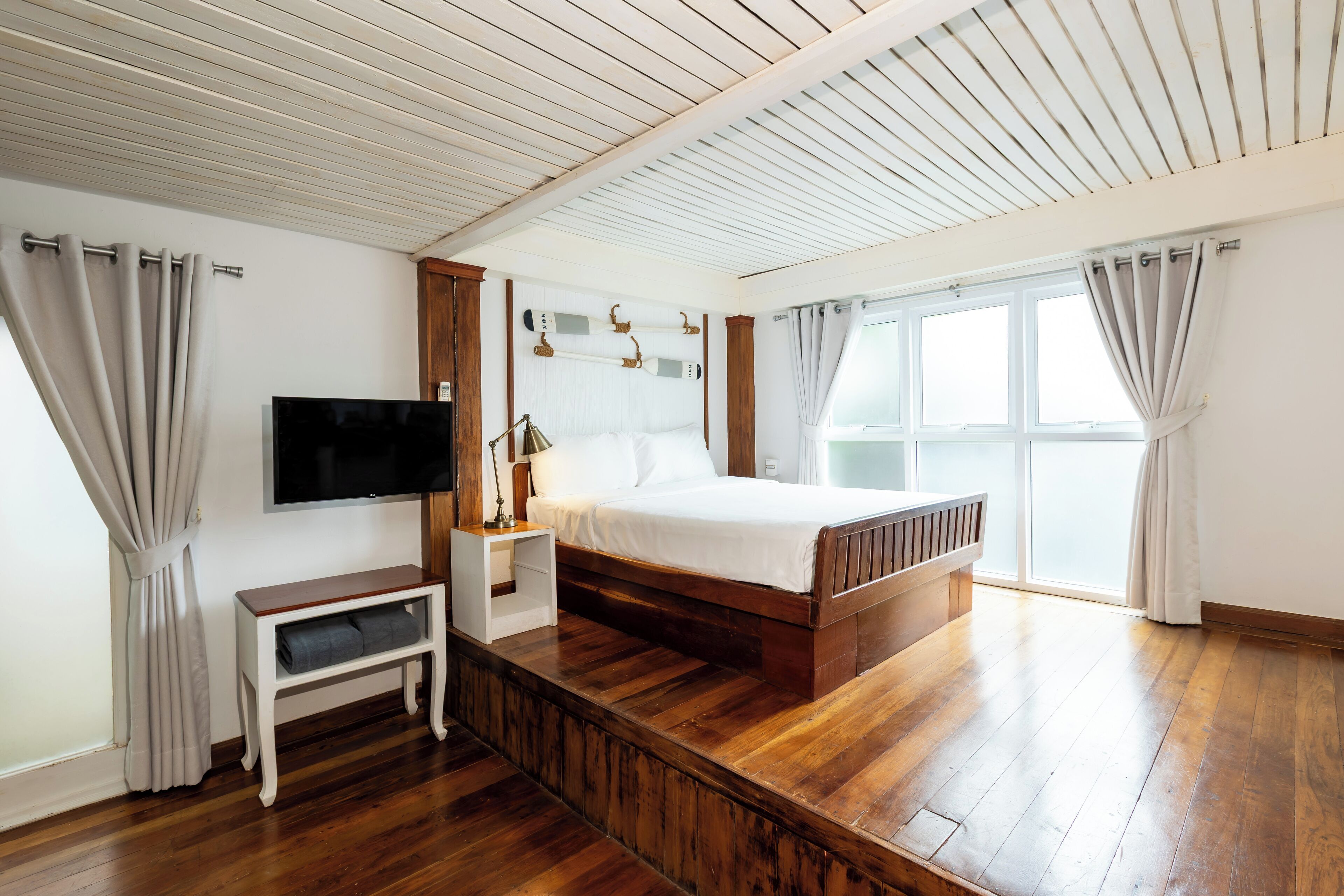 A bedroom with a wooden bed, a TV mounted on the wall, a bedside table, and a window with curtains.