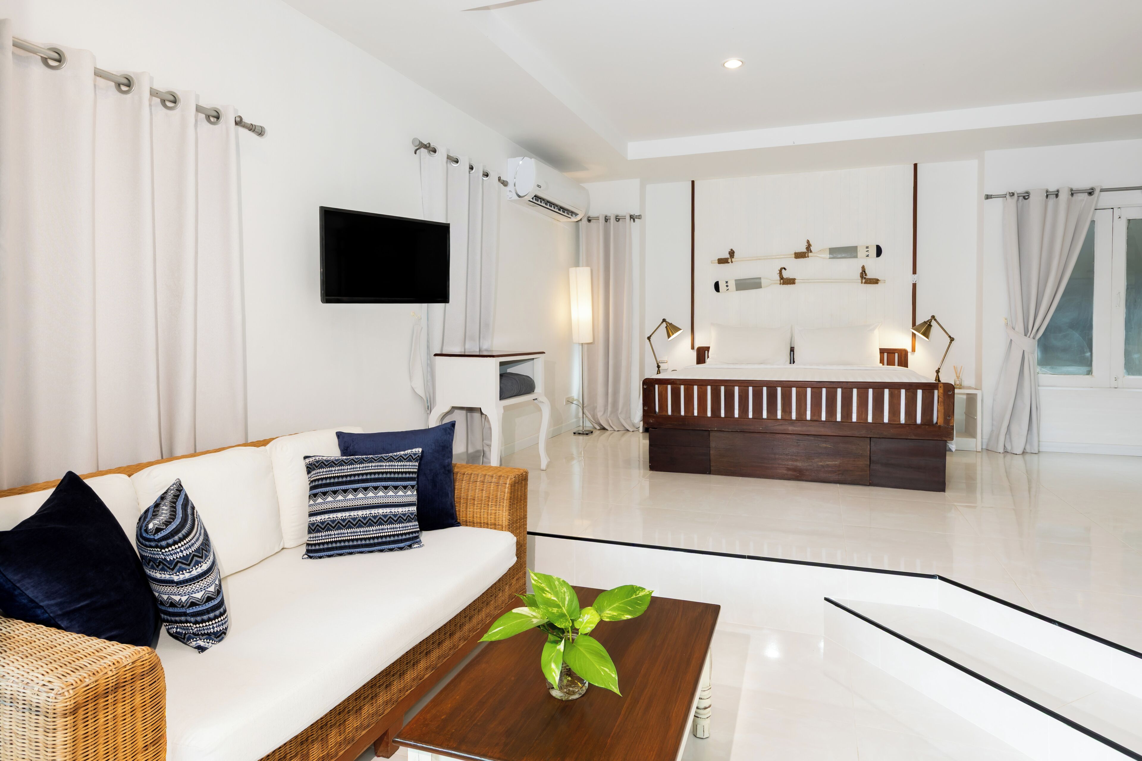 A modern hotel room with a white sofa, a wooden coffee table, a flat-screen TV, a large mirror, and a bathroom with a shower and toilet.
