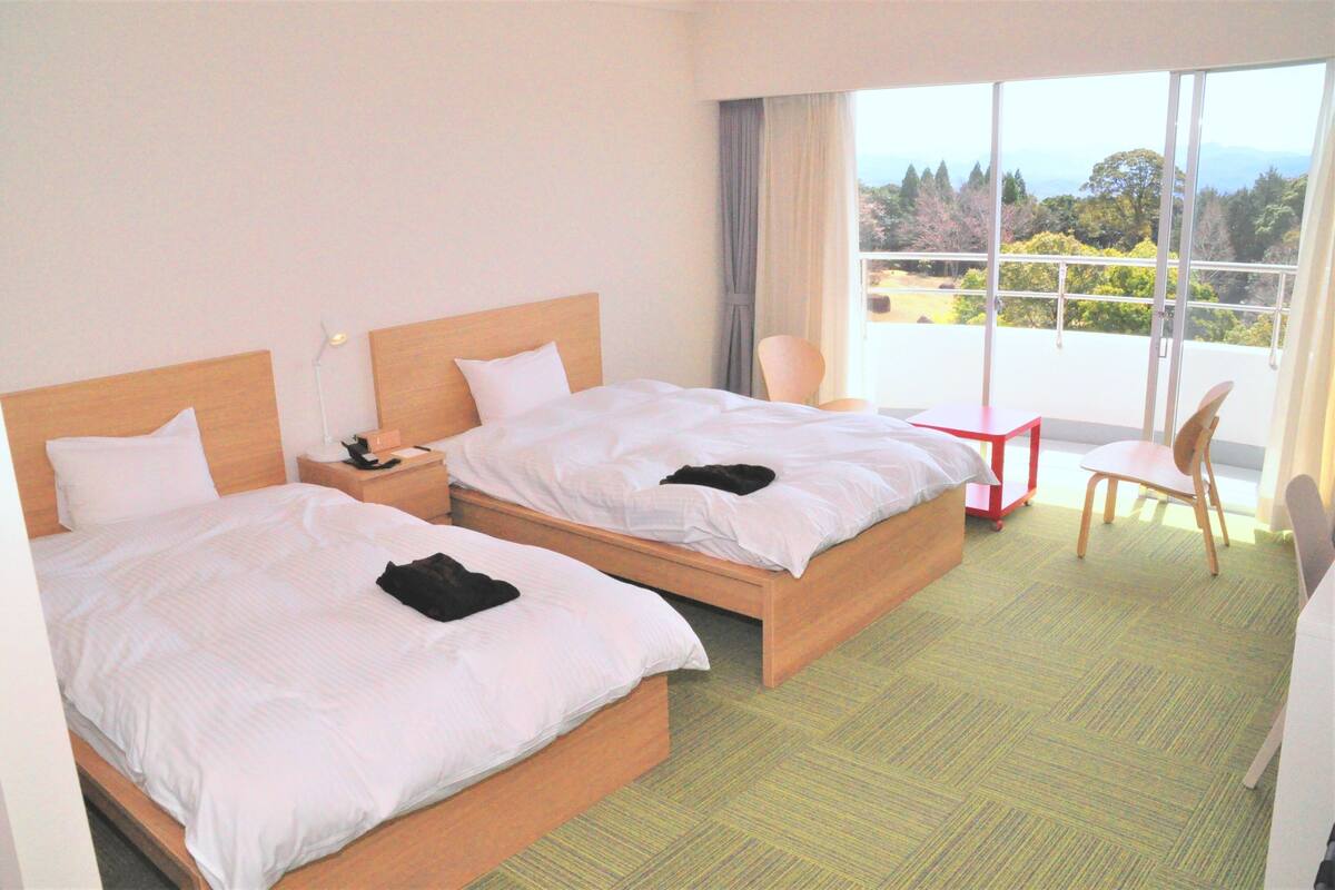 A hotel room with two beds, a small table, a chair, and a large window offering a view of the outdoors.