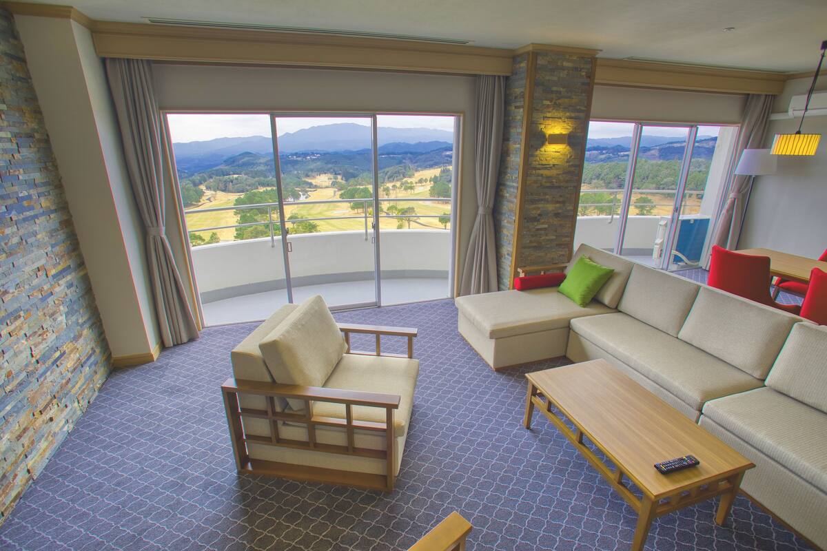 A room with a sofa, a chair, a small table, and a view of a landscape through large windows.