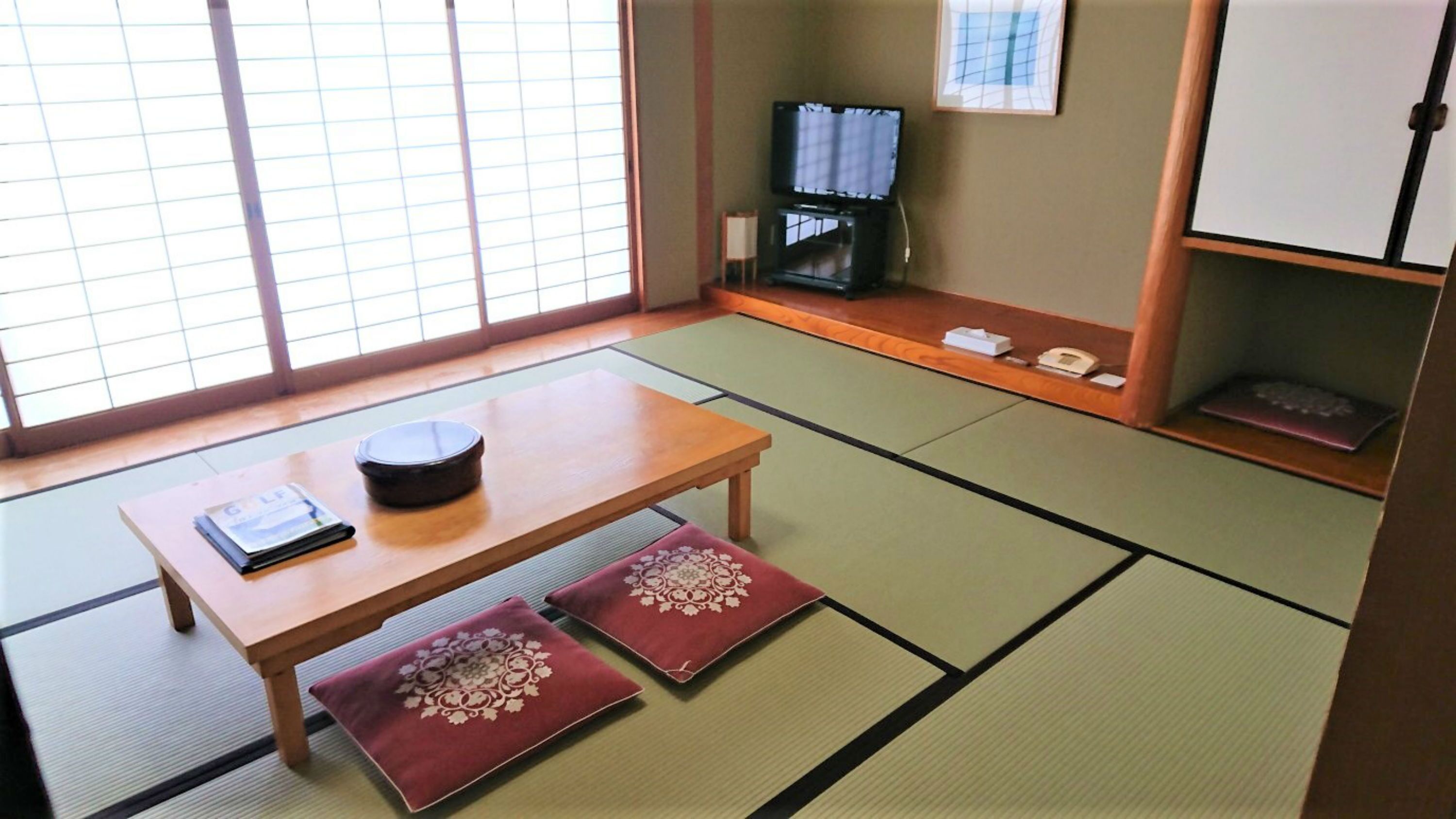 A traditional Japanese room with tatami flooring, a low wooden table, two cushions, a television, and a cabinet.