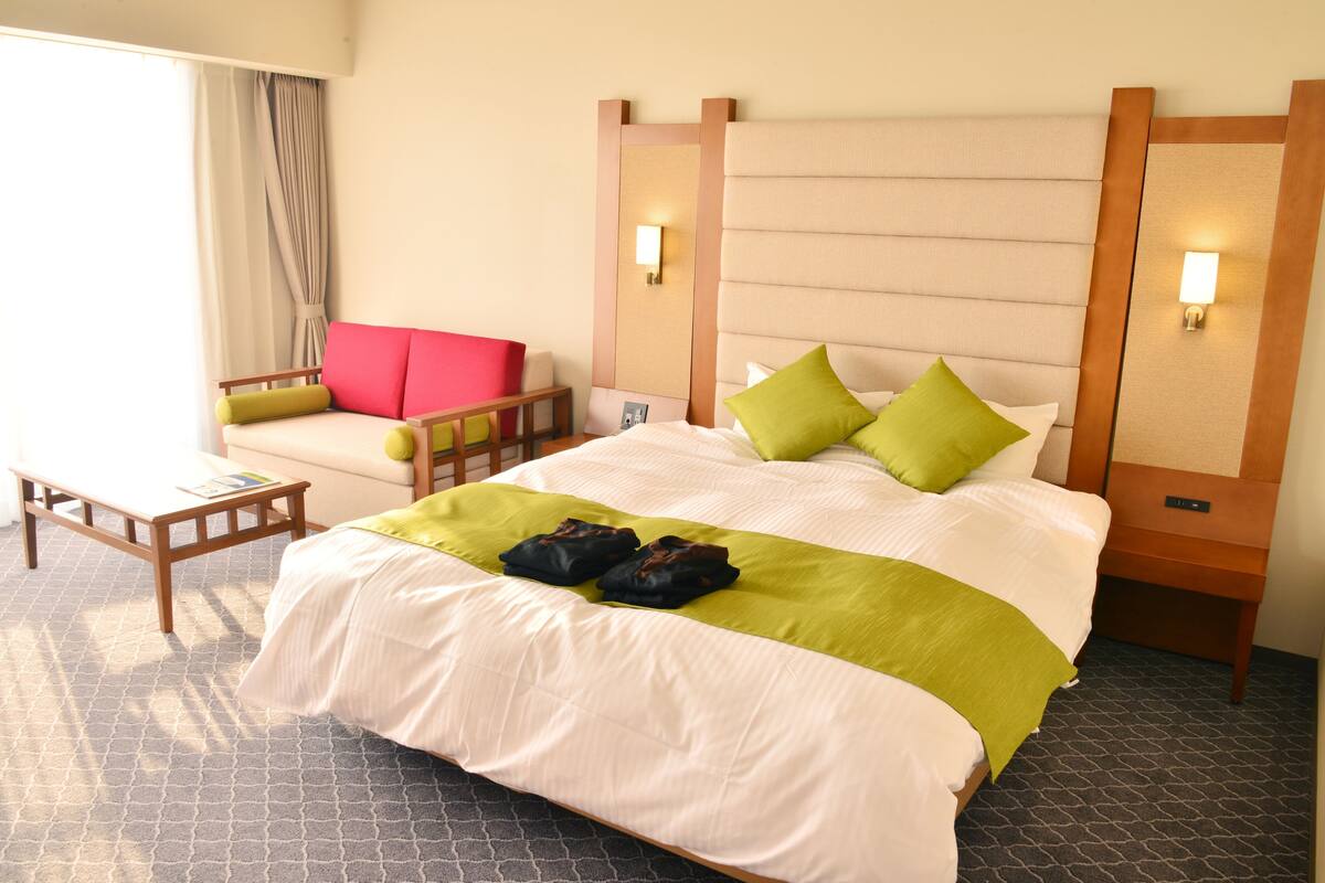 A hotel room with a large bed, a wooden nightstand, a bedside table, a chair, and a window with curtains.