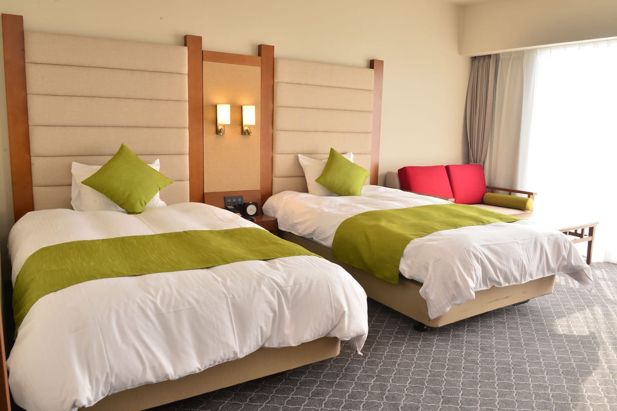 A hotel room with two beds, each with white bedding and green accents, a red chair, and a nightstand with a clock.