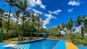 Piscina com deck azul e amarelo, rodeada por palmeiras e vegetação.