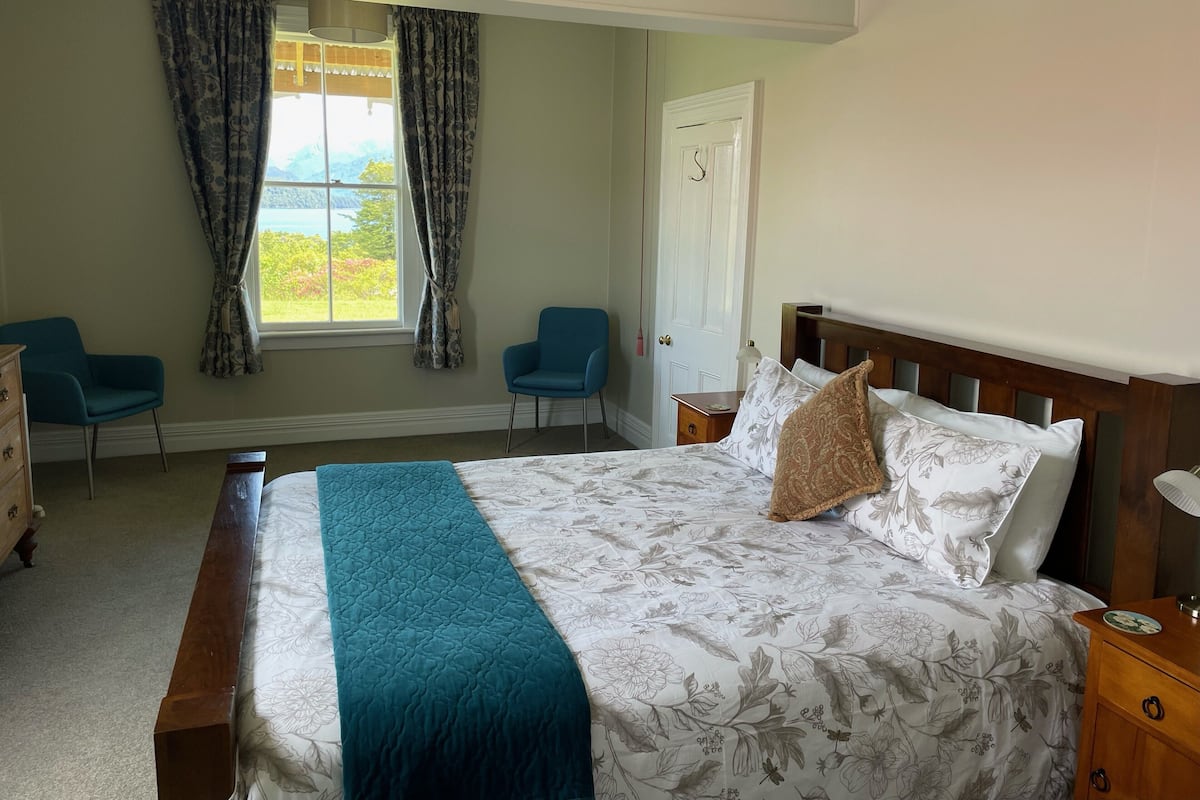 A bedroom with a bed, two chairs, a nightstand, a door, and a window with curtains.