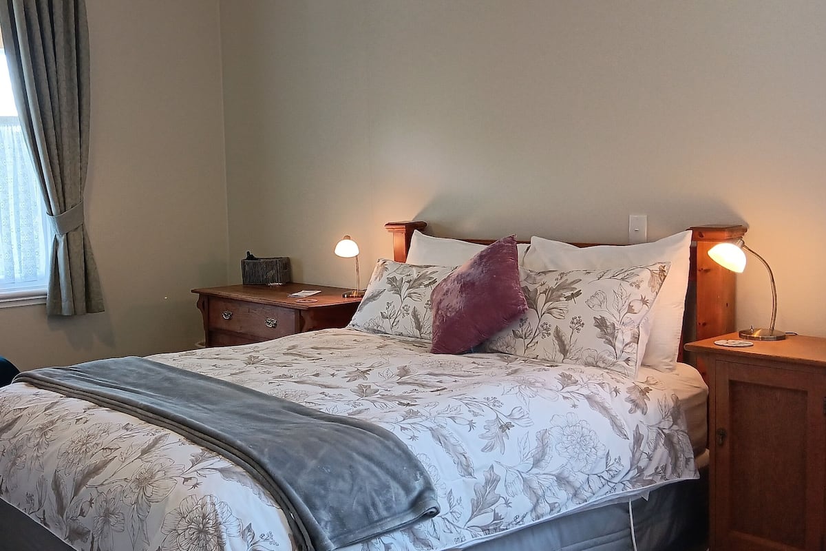 A bedroom with a bed, bedside tables, a nightstand, a window with curtains, and a small bedside lamp.