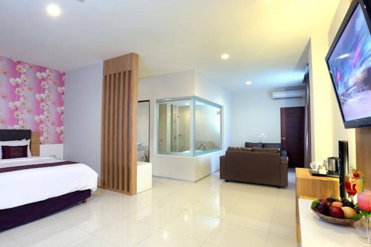 Una habitación de hotel moderna con una cama grande, un baño con cabina de ducha de vidrio, una sala de estar con sofá y un televisor montado en la pared.