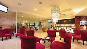 A hotel lobby with red velvet chairs arranged around a central table with a teapot and cups. A large, ornate light fixture hangs from the ceiling, and a television is mounted on the wall. - Lombok Plaza Hotel & Convention (Mataram)