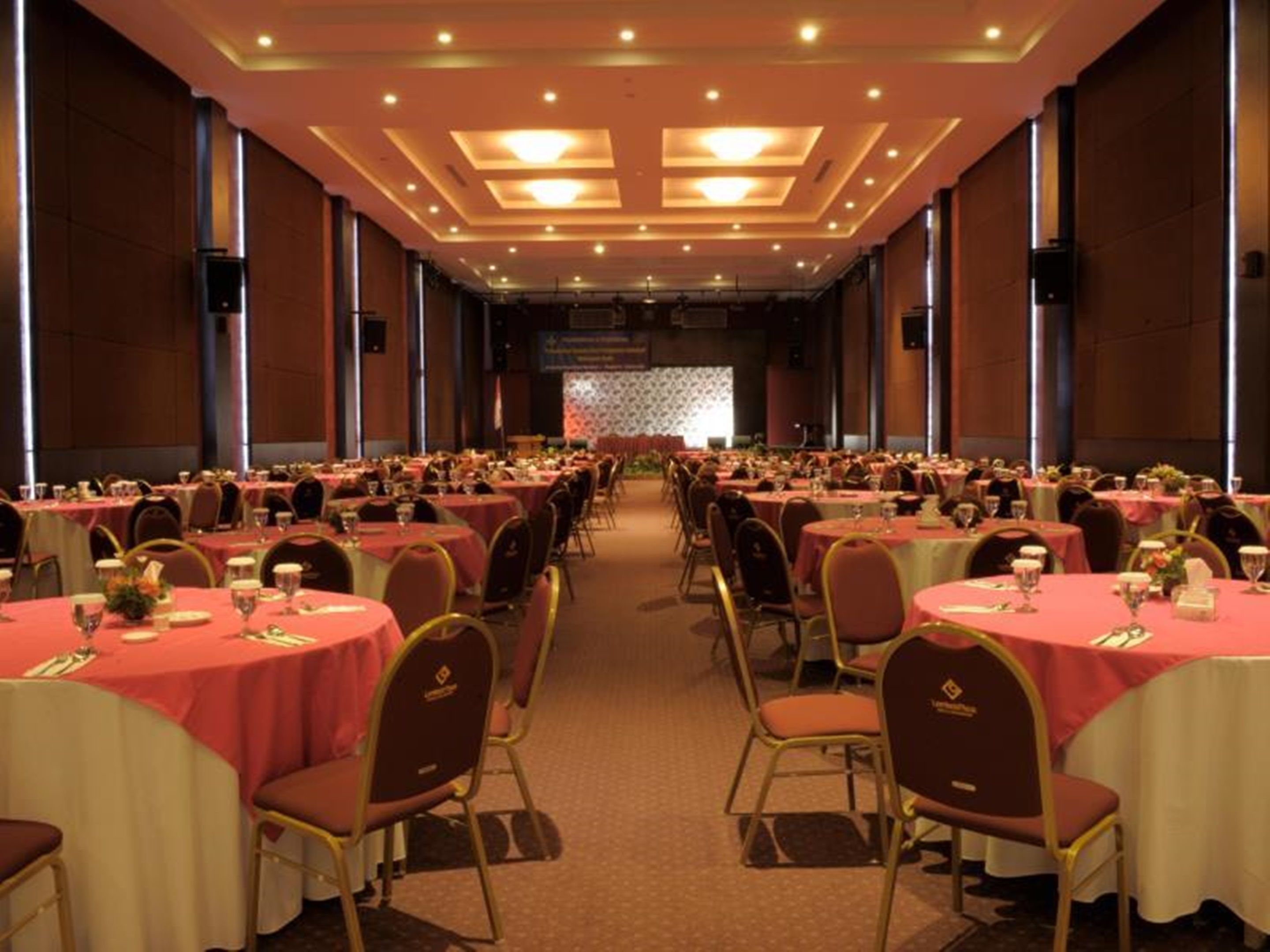 A large banquet hall with rows of round tables covered in red tablecloths, arranged in a U-shape facing a stage.