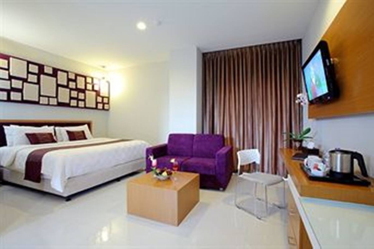 Una habitación de hotel moderna con una cama, un sofá morado, una mesa de centro, un televisor montado en la pared y una ventana con cortinas.