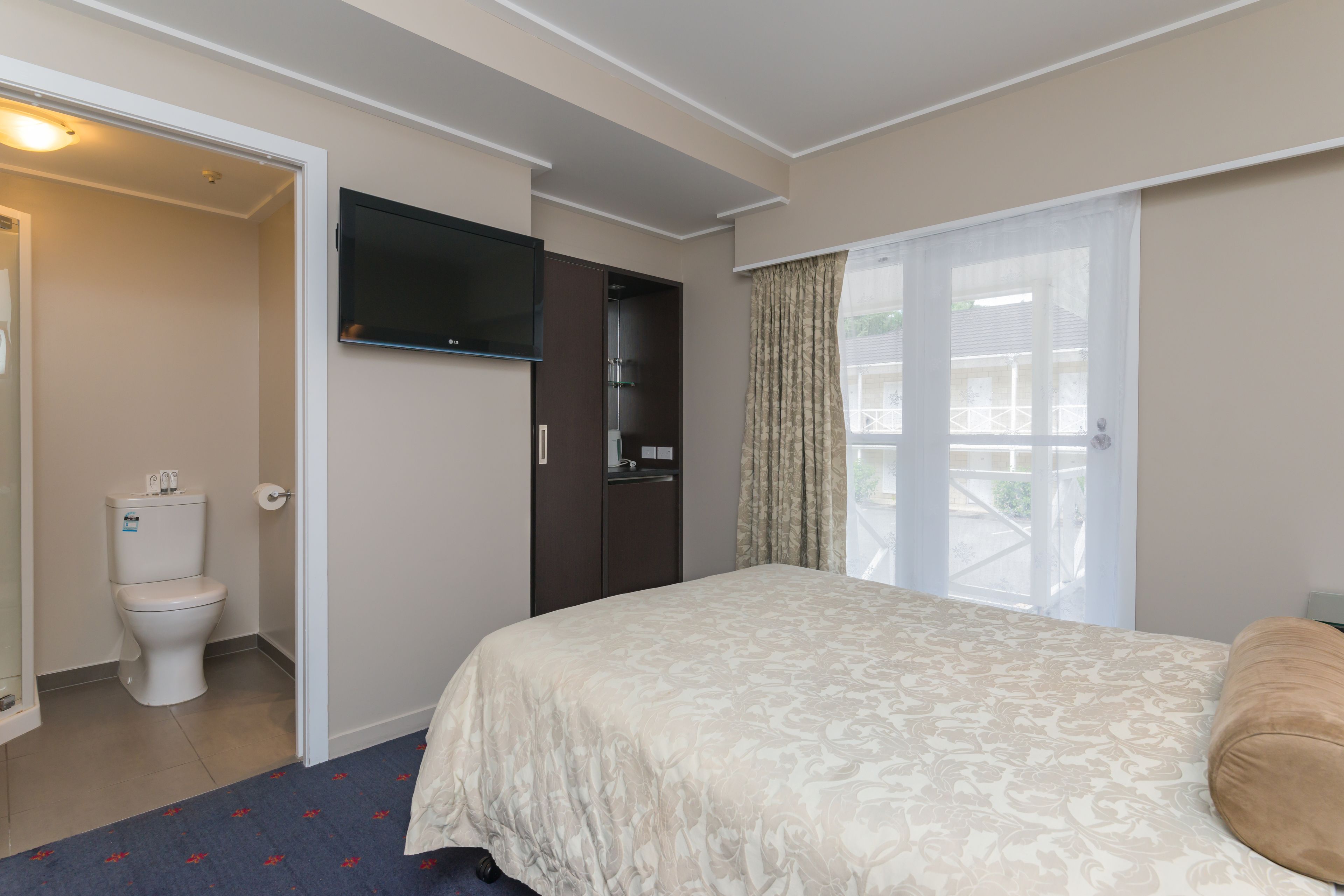 A hotel room with a bed, a television mounted on the wall, a bathroom with a toilet and sink, and a window with curtains.