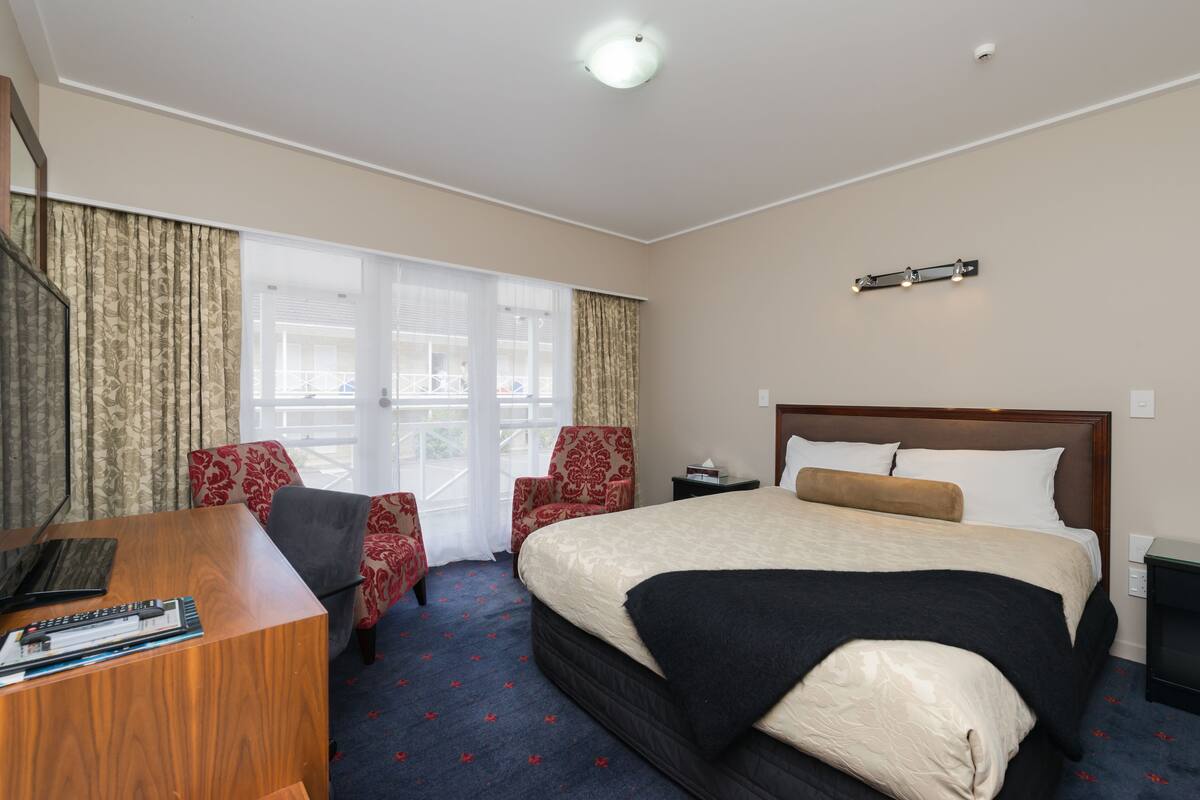 Ein Hotelzimmer mit einem großen Bett, einem Schreibtisch mit einem Laptop, zwei Sesseln und Blick durch große Fenster nach draußen.