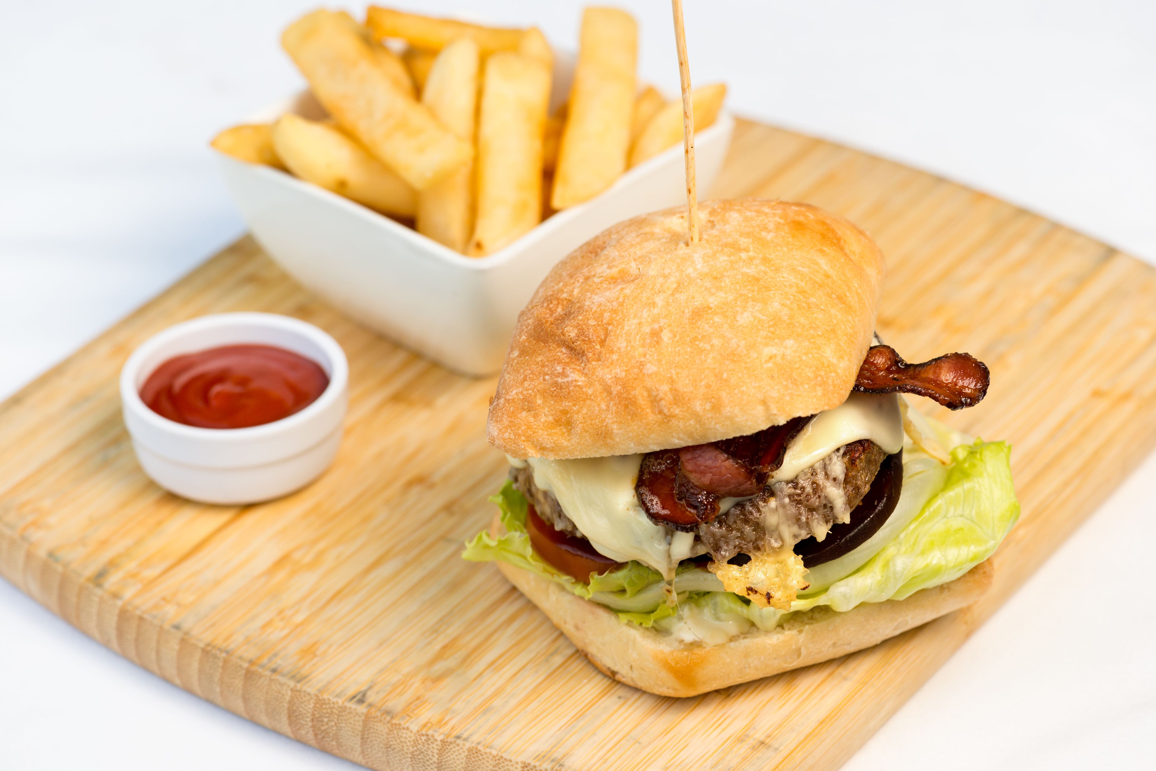 Un hamburger garni de bacon, de laitue, de tomates et de fromage, servi dans un pain, accompagné d’une portion de frites et d’un petit bol de ketchup.