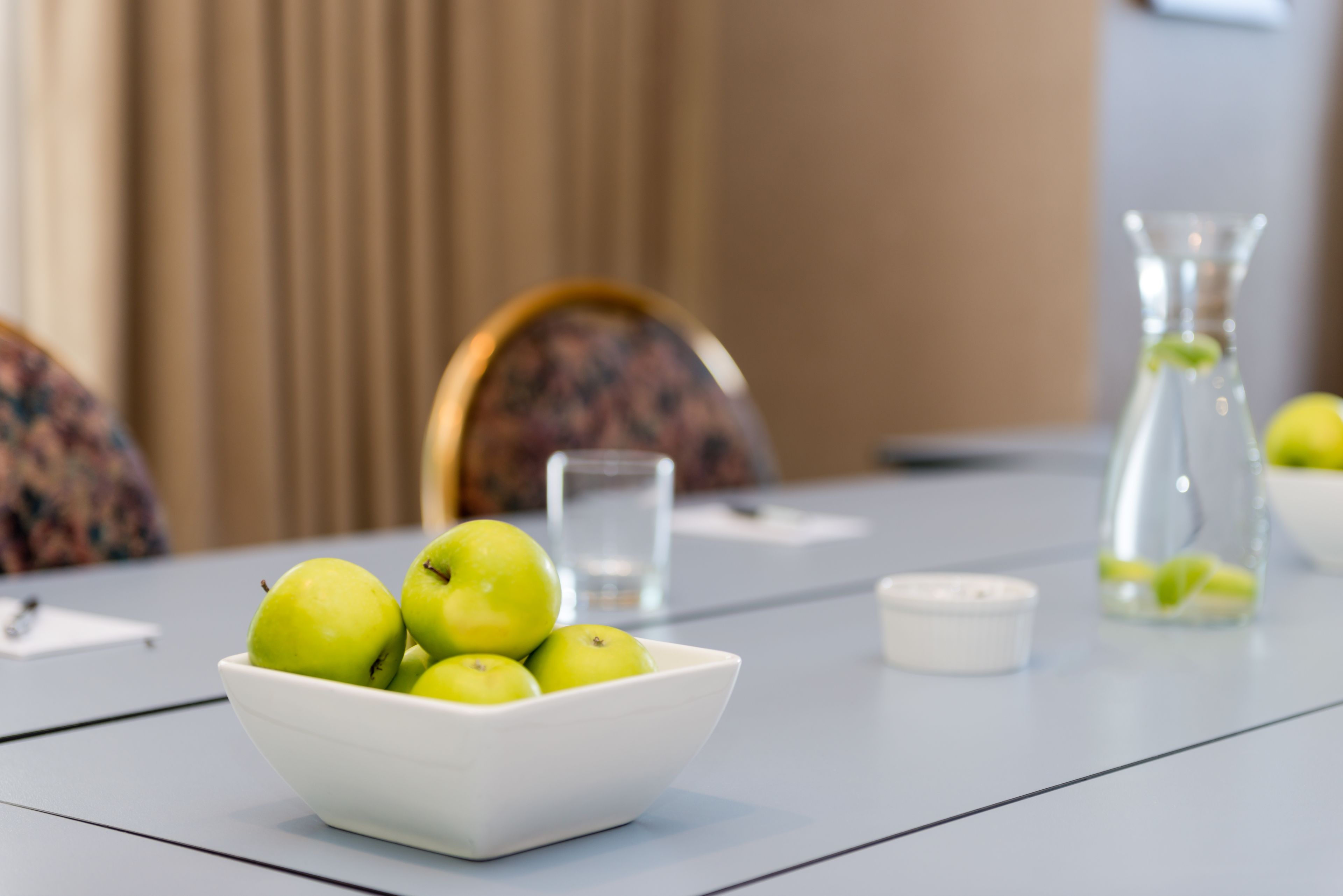A white bowl of green apples on a table with a glass of water, a candle, and a glass bottle in the background.