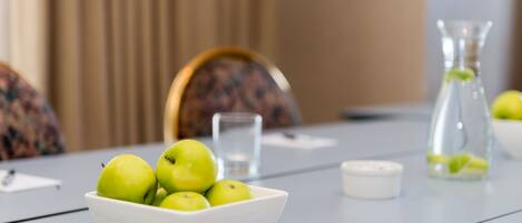 A white bowl of green apples on a table with a glass of water, a candle, and a glass bottle in the background.