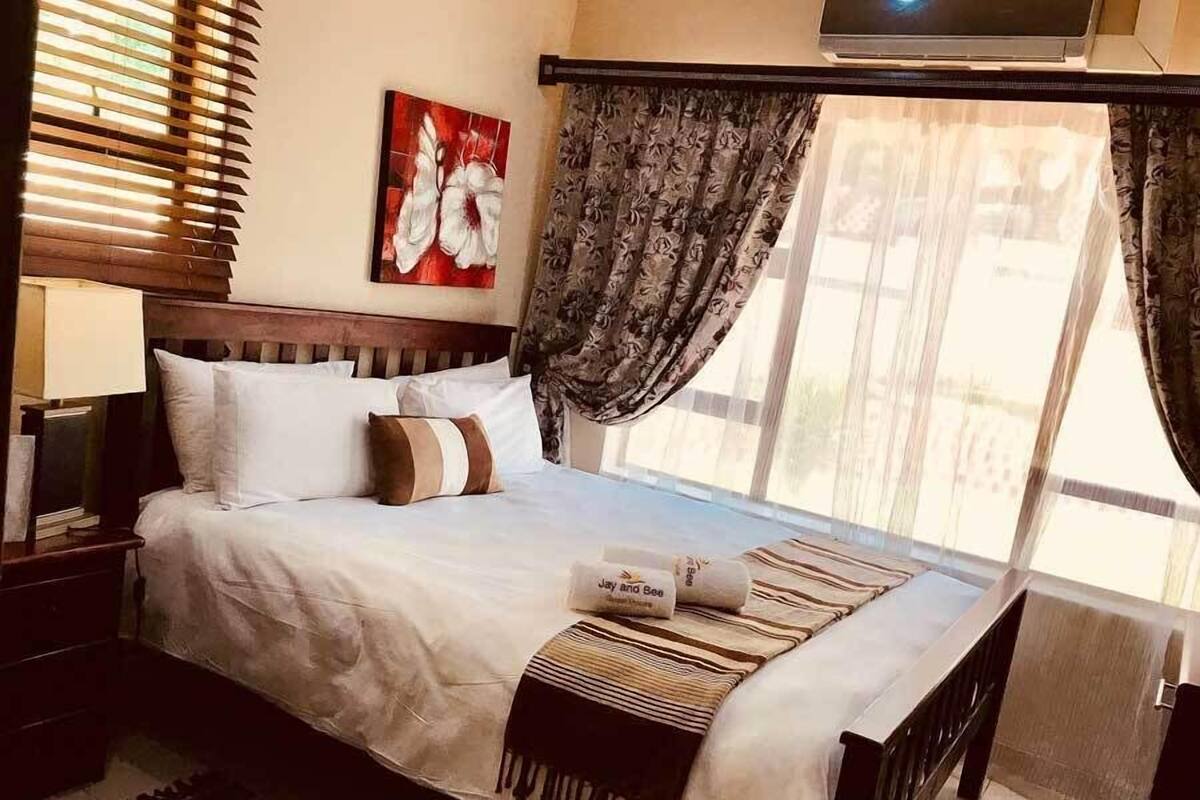 Una cama bien hecha con ropa blanca y un plaid a rayas, un cabecero de madera, un cuadro en la pared y una ventana con cortinas.