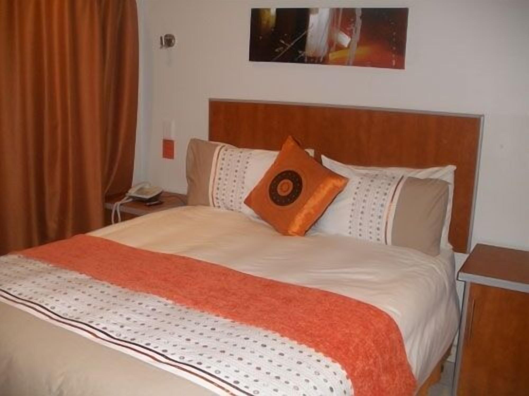 A neatly made bed with a patterned bedspread, a single orange pillow, and a wooden headboard.