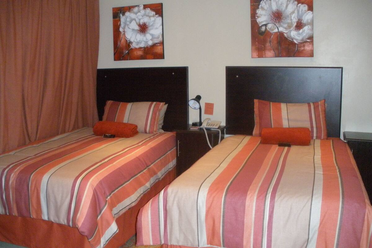 Dos camas dobles con ropa de cama a rayas, una mesita de noche con lámpara, y dos cuadros de flores colgados en la pared.