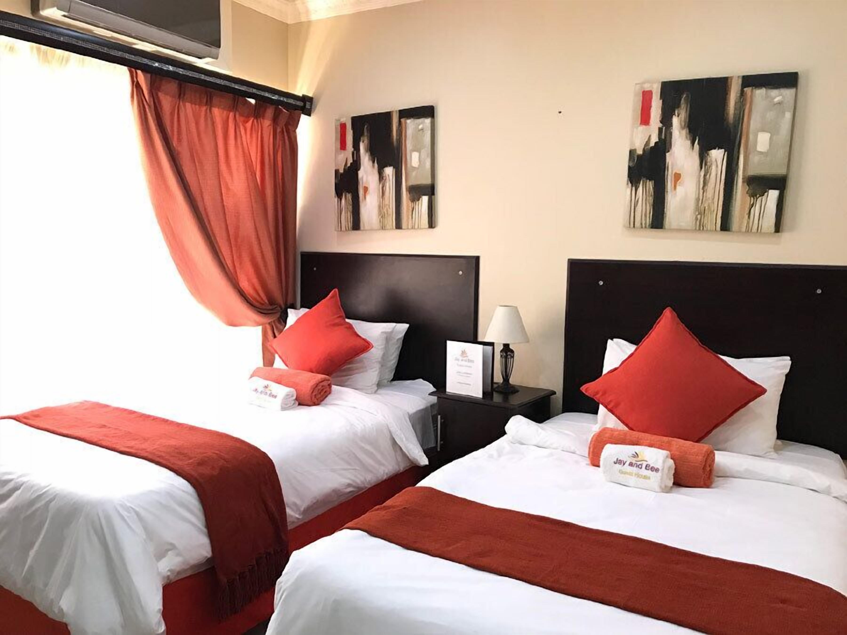 A hotel room with two beds, each with white bedding and red pillows. There are two nightstands with lamps and a sign on one. The room has a large window with curtains, and artwork is displayed on the walls.