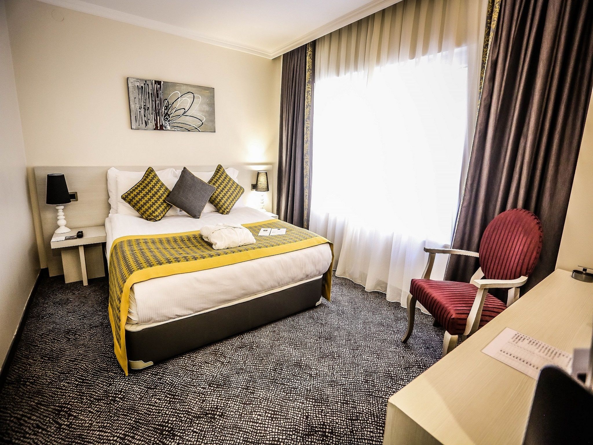 Quarto de hotel com cama, escrivaninha, cadeira, mesa de cabeceira com abajur e janela com cortinas.