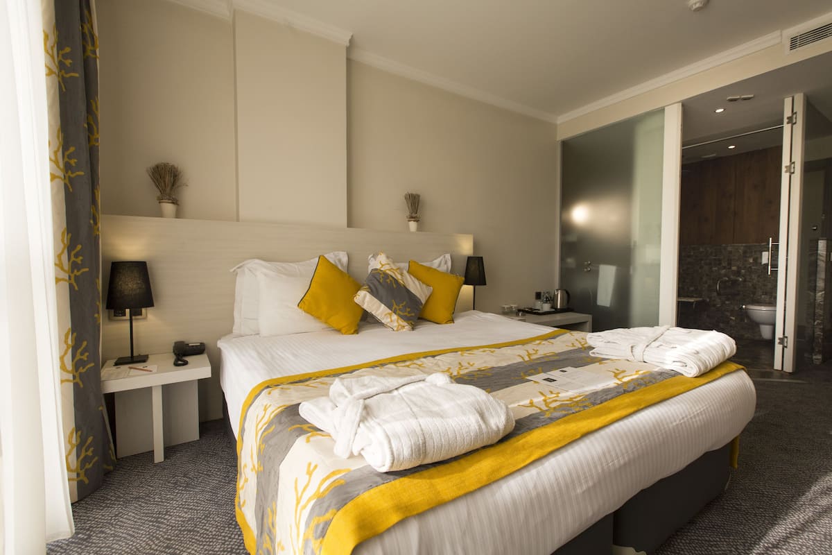 A hotel room with a large bed, bedside tables, a nightstand, a bathroom visible through an open door, and a shower area.