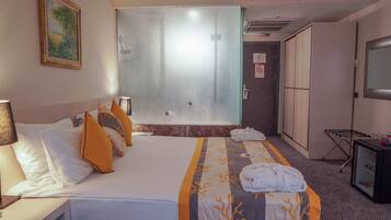 Habitación de hotel con cama, mesita de noche, lámpara, bata, toalla, televisor y puerta con espejo.