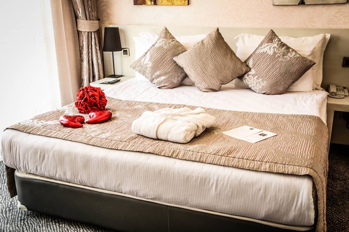 A neatly made bed with a beige bedspread, a red rose, and a white towel. There are four pillows on the bed, and a nightstand with a lamp and phone is visible to the right.