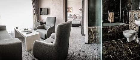 Habitación de hotel con zona de estar, televisión, cama y baño con ducha y lavamanos.