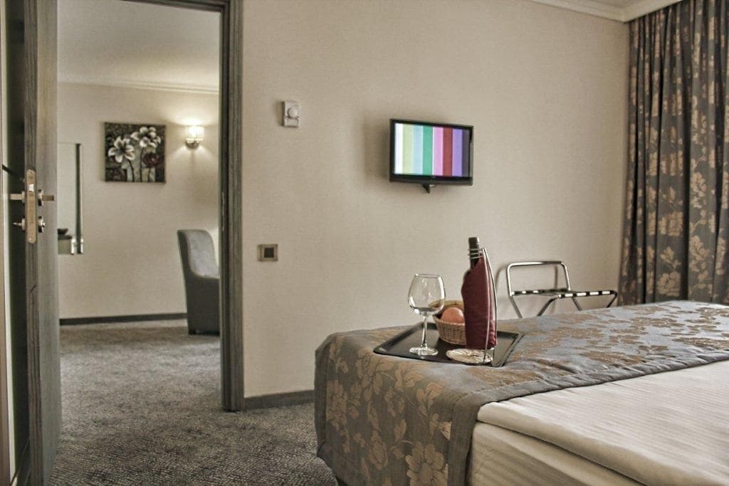 Habitación de hotel con cama, mesita de noche con botella y vaso, televisor en la pared y una silla.