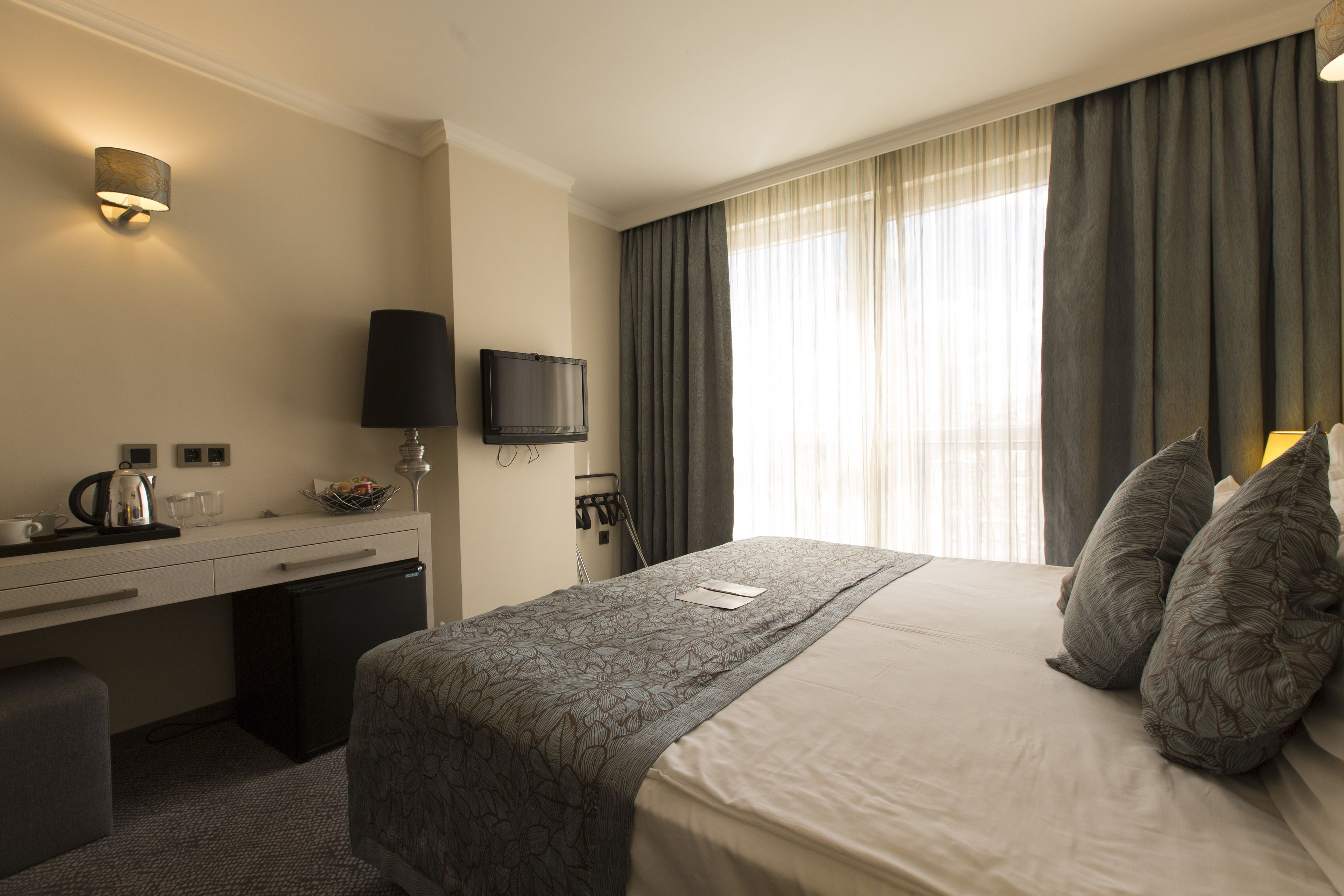 Una habitación de hotel con cama, escritorio, televisor, cafetera y ventana con cortinas.