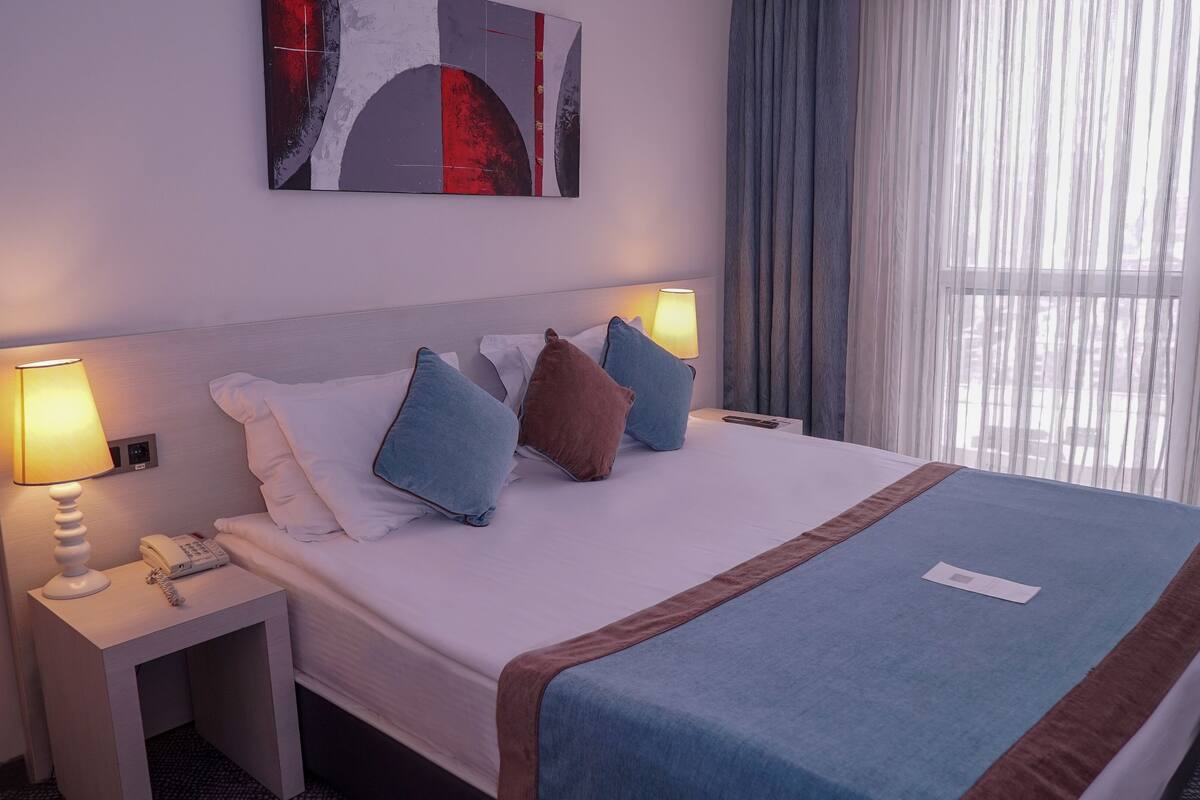 A hotel room with a large bed, two bedside tables, a lamp, a painting, and a window with curtains.