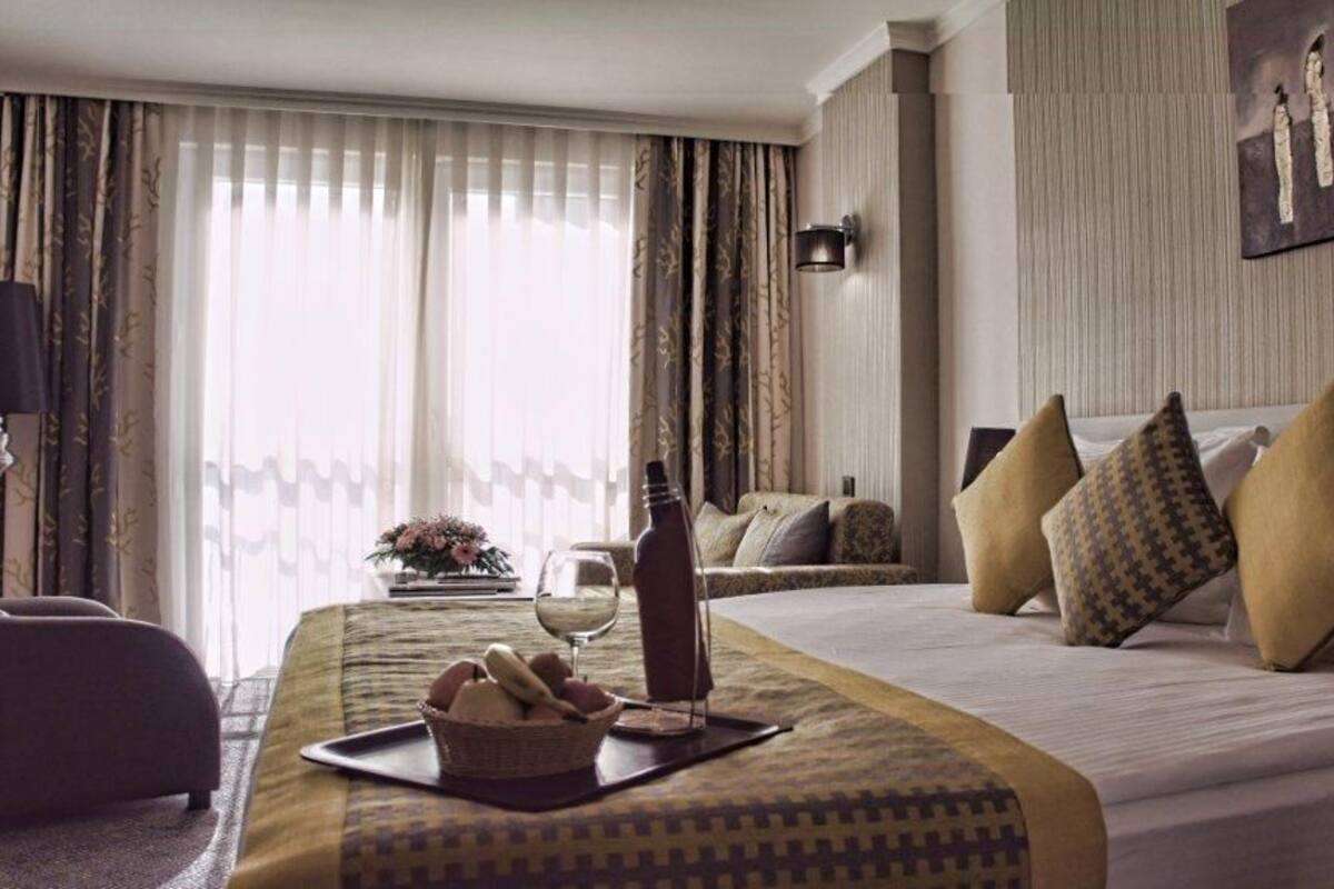 A hotel room with a large bed, a bedside table with a wine glass, a basket of bread, and a view of the outdoors through sheer curtains.
