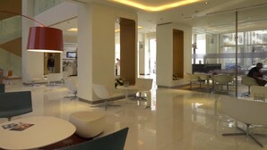 A modern hotel lobby with a spacious layout, featuring a central seating area with round tables and chairs, a red pendant light, and a glass partition separating the dining area from the lobby. - Ibis Yanbu Saudi Arabia (Yanbu)
