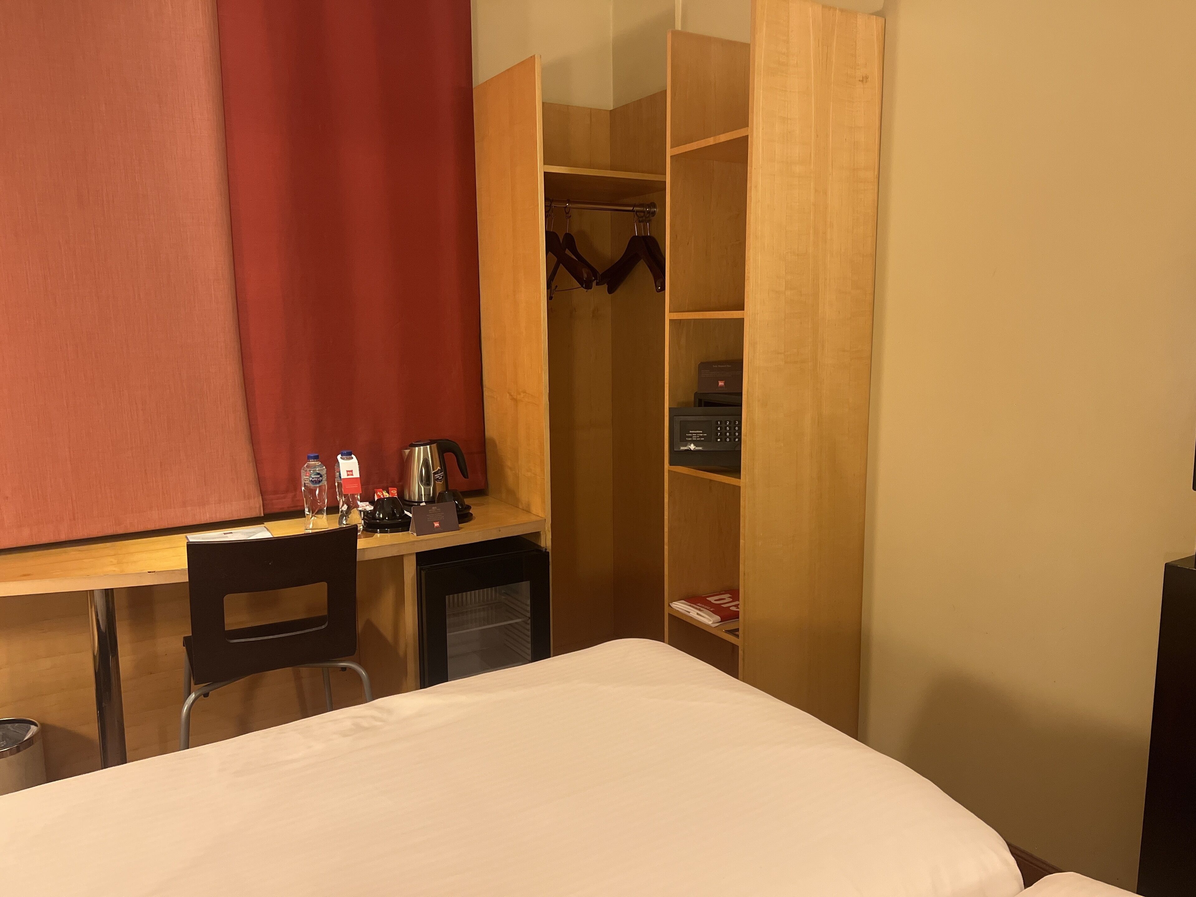 A hotel room with a bed, a desk with a chair, a wardrobe, and a red curtain.