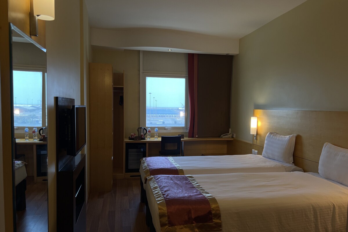A hotel room with two beds, a desk with a chair, a TV, and a window with a view of the airport.