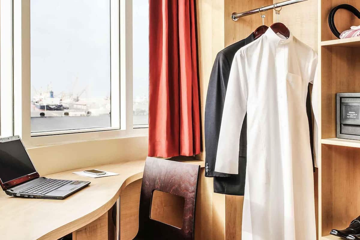 A hotel room with a wooden desk, a laptop, a chair, a coat rack with a coat, and a view of a marina through the window.