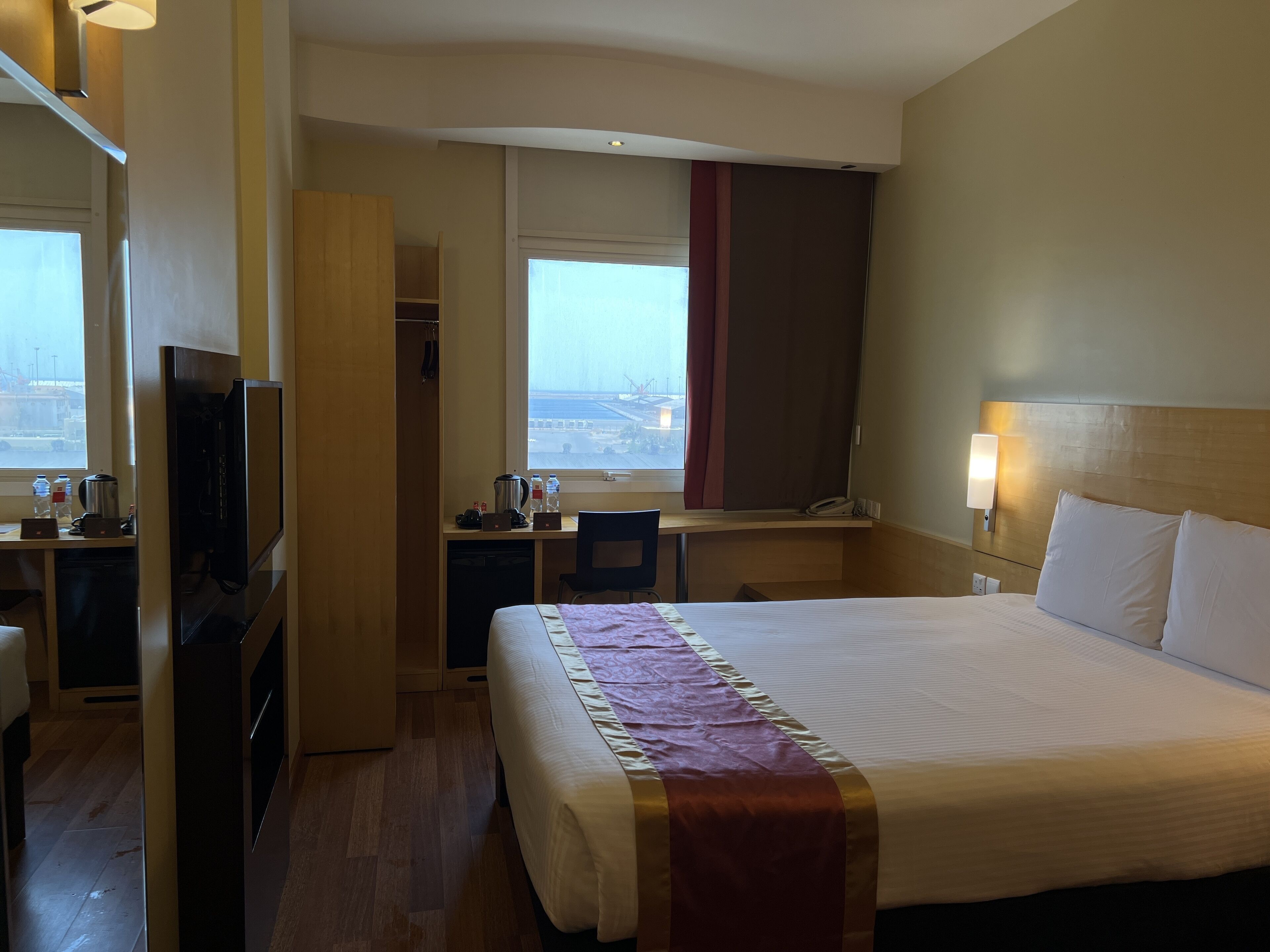 A hotel room with a bed, a desk, a television, a wardrobe, and a window with a view of the city.