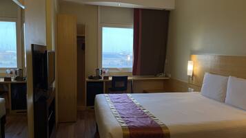 A hotel room with a bed, a desk, a television, a wardrobe, and a window with a view of the city.