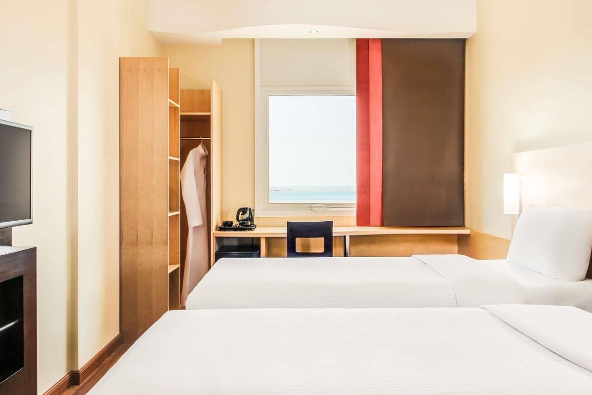 A hotel room with a bed, a desk, a TV, and a view of the sea.