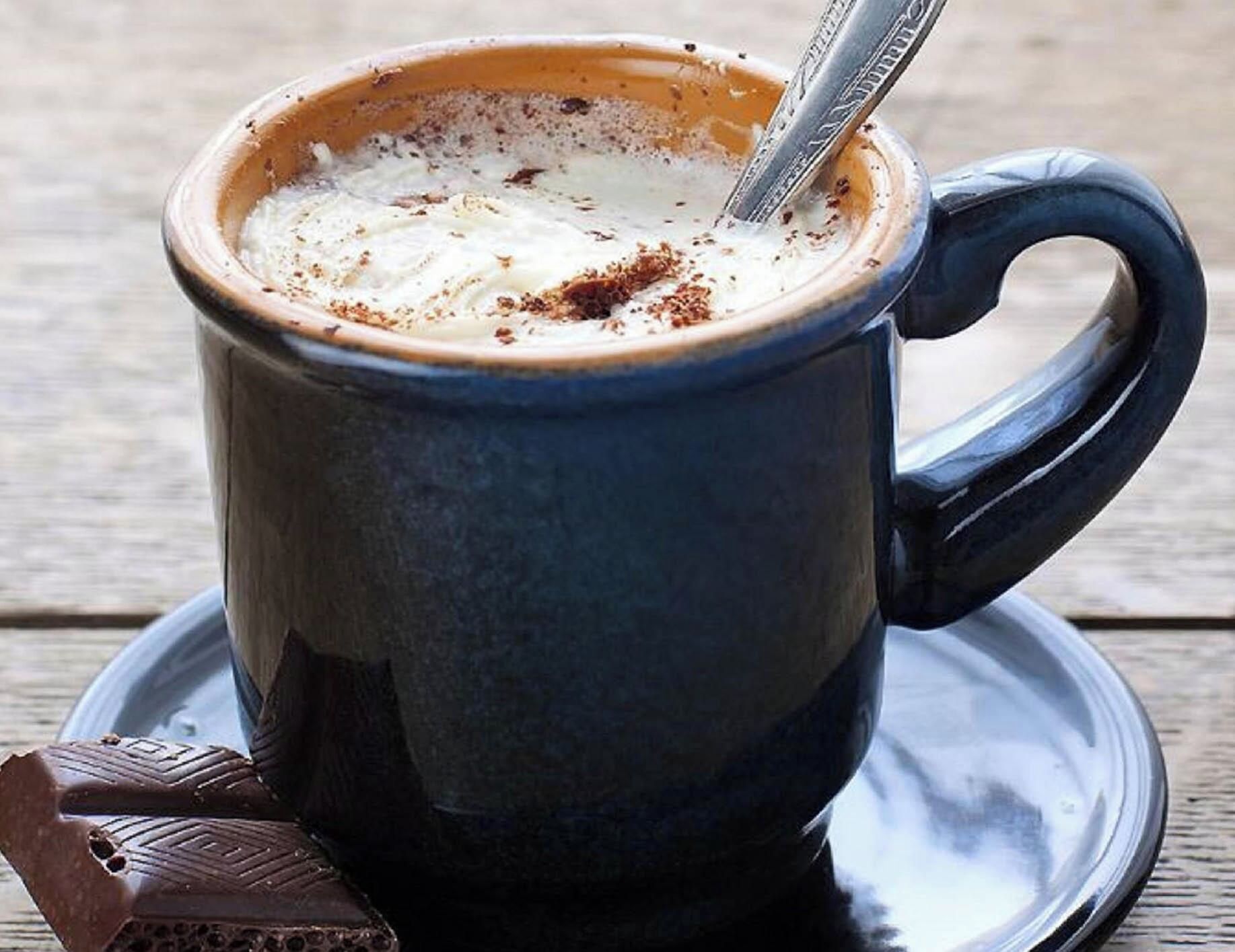 A dark blue cup with a handle, filled with a frothy beverage, possibly coffee or hot chocolate, and a spoon inside.