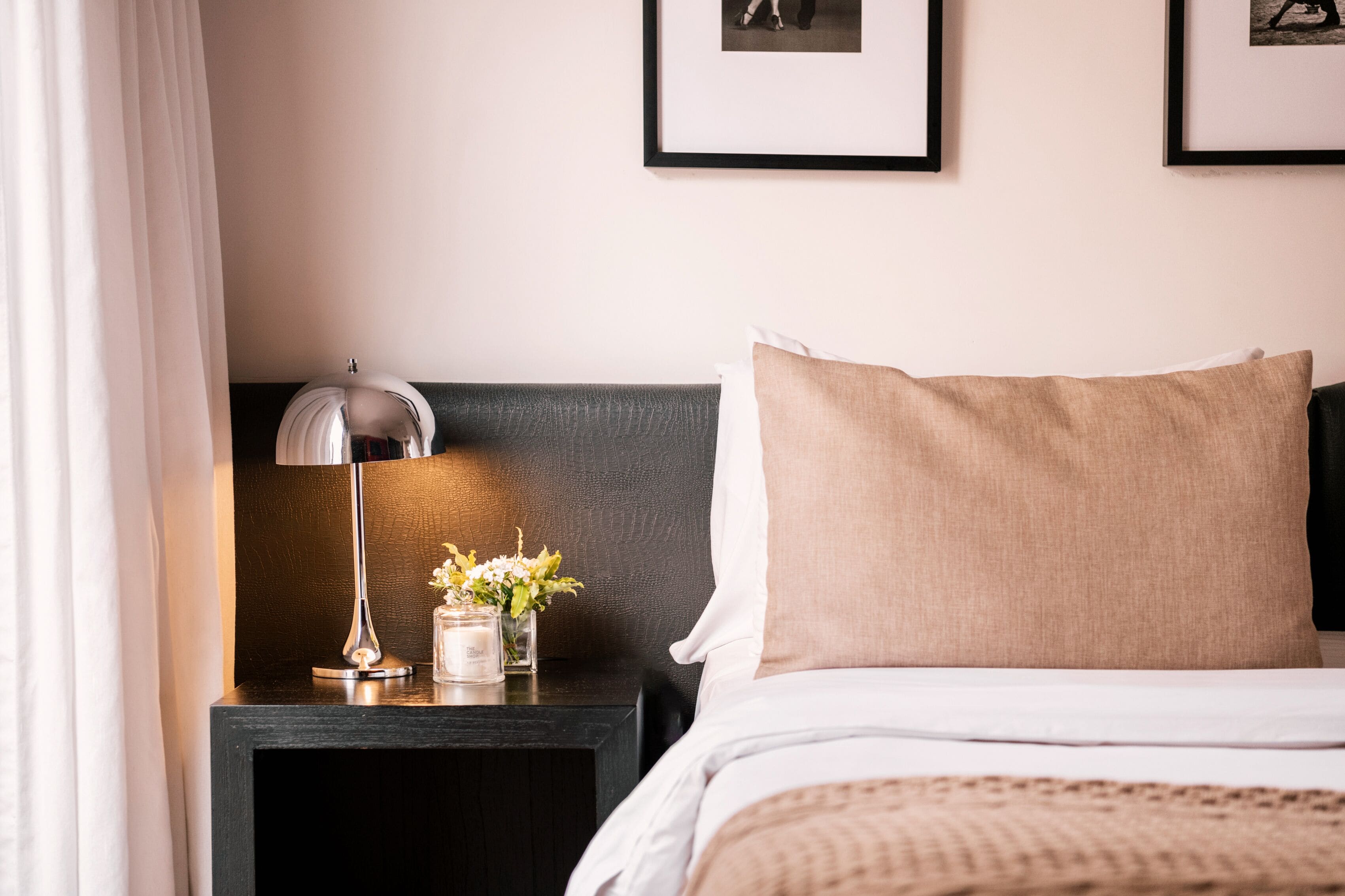 Una cama bien hecha con un almohadón color beige y una manta blanca, una mesita de noche con una lámpara y un florero, y dos cuadros en la pared.