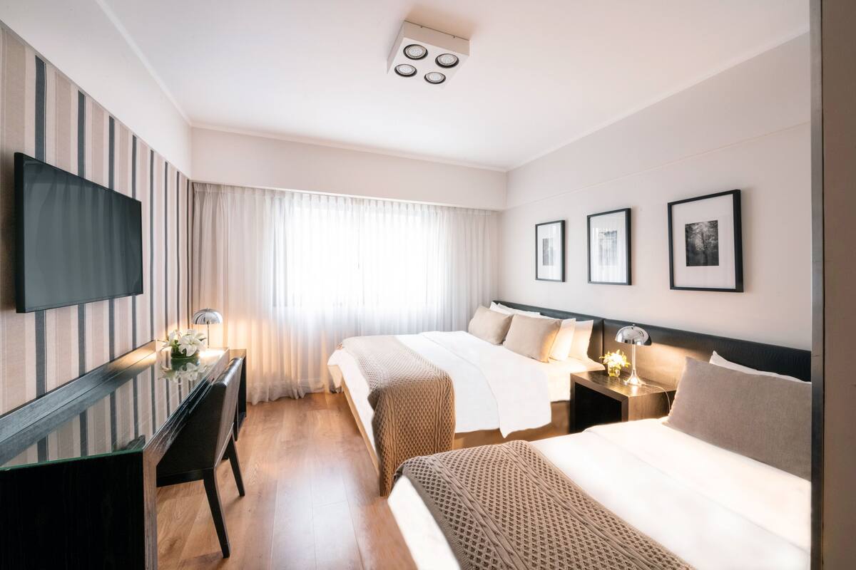 Habitación de hotel con una cama grande, una cama más pequeña, un escritorio con silla, televisor montado en la pared y tres cuadros enmarcados.