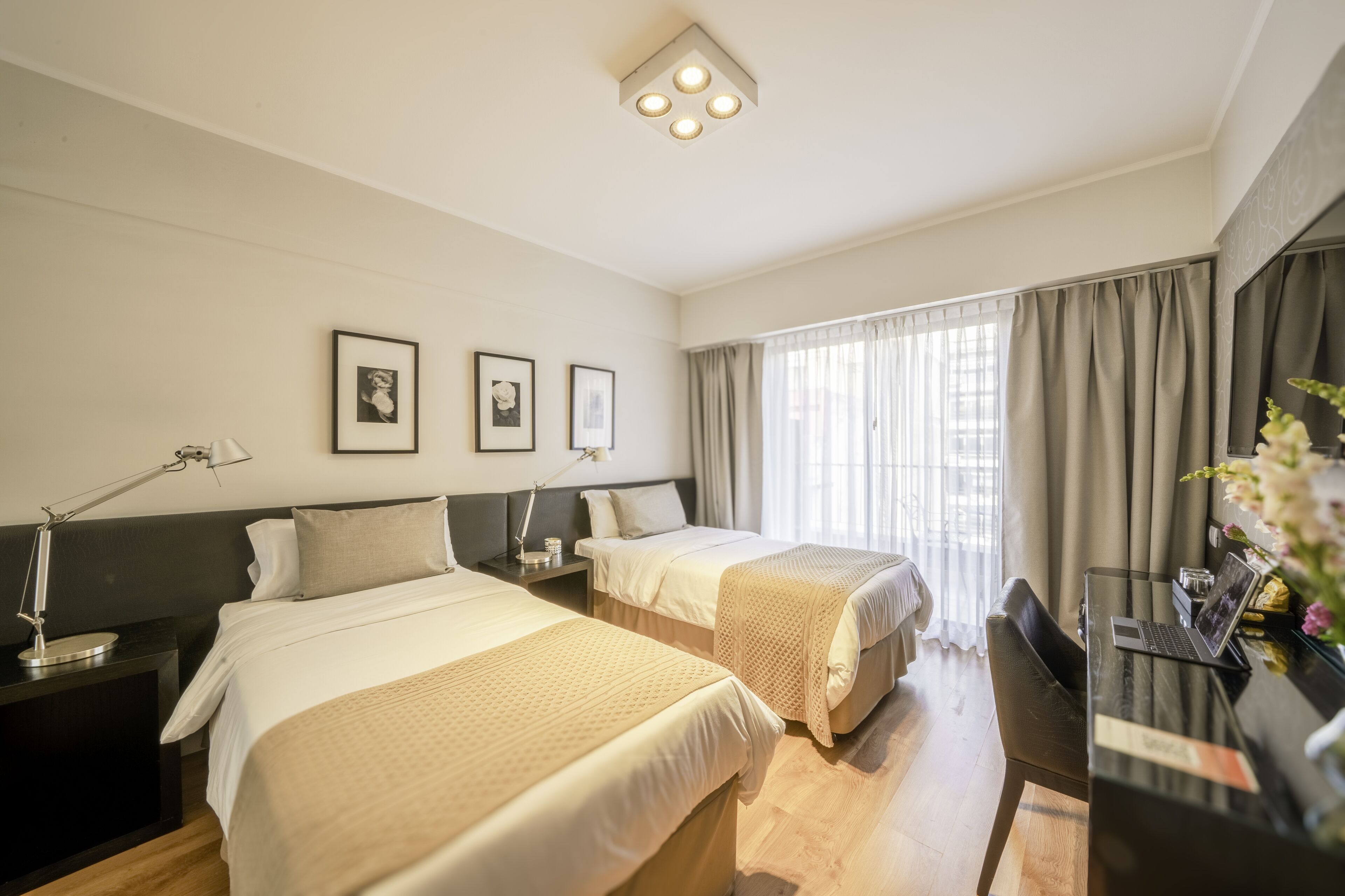 A hotel room with two beds, a desk with a laptop, a desk lamp, and a large window with curtains.